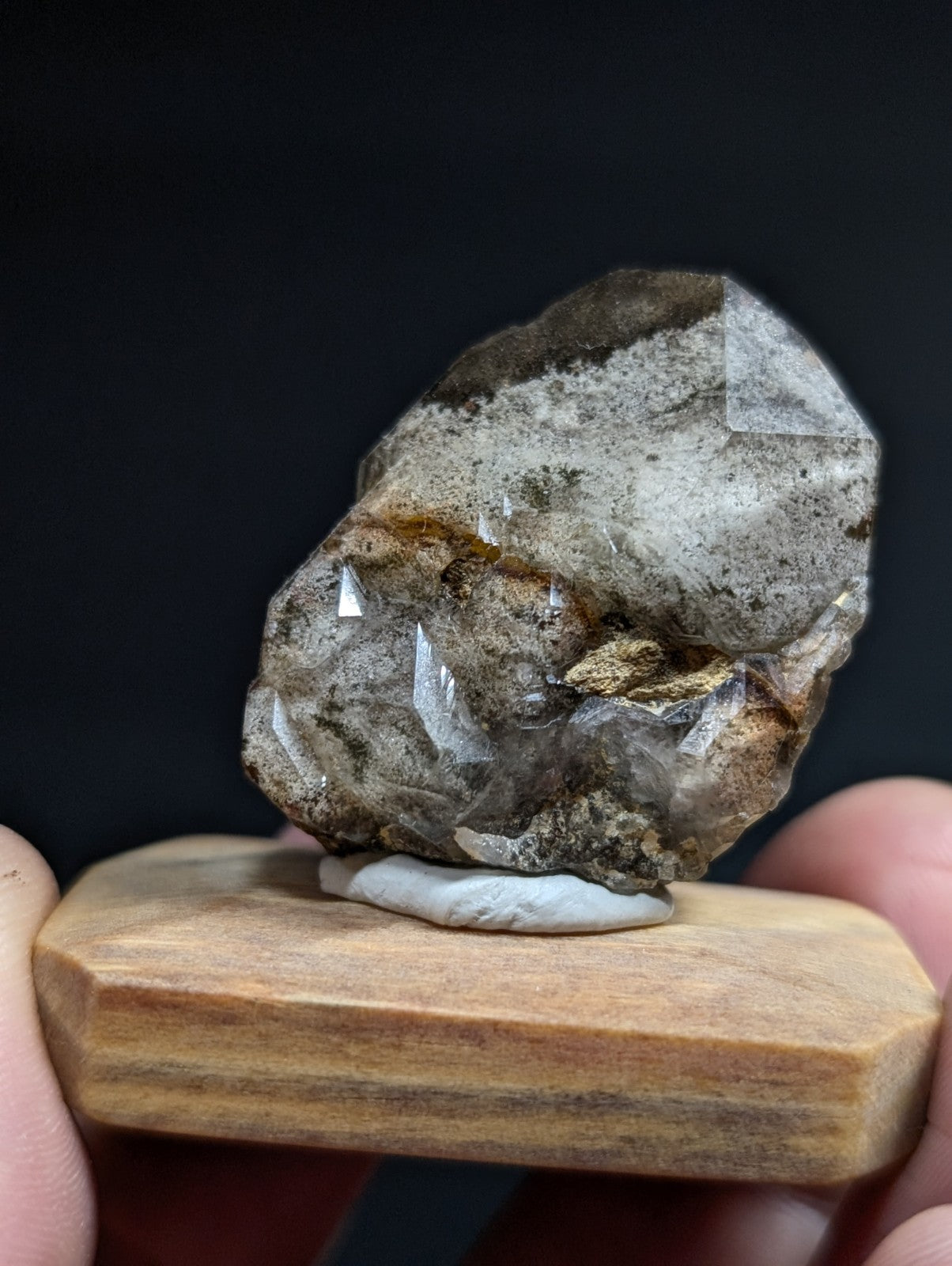 A translucent, faceted quartz crystal with earthy brown and gray veining rests on a wooden display stand.