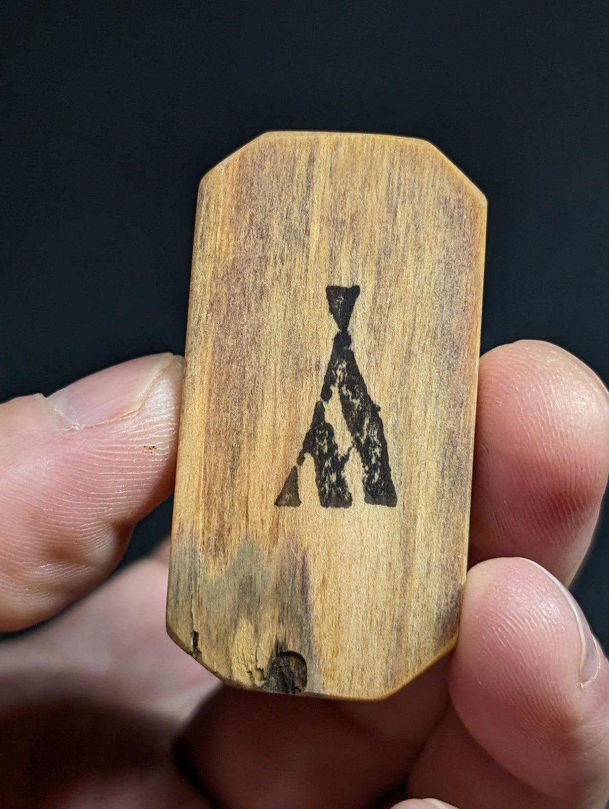 A hand holding a light brown wooden rectangular tag with rounded corners, featuring a black burned-in silhouette of an a-frame tent.