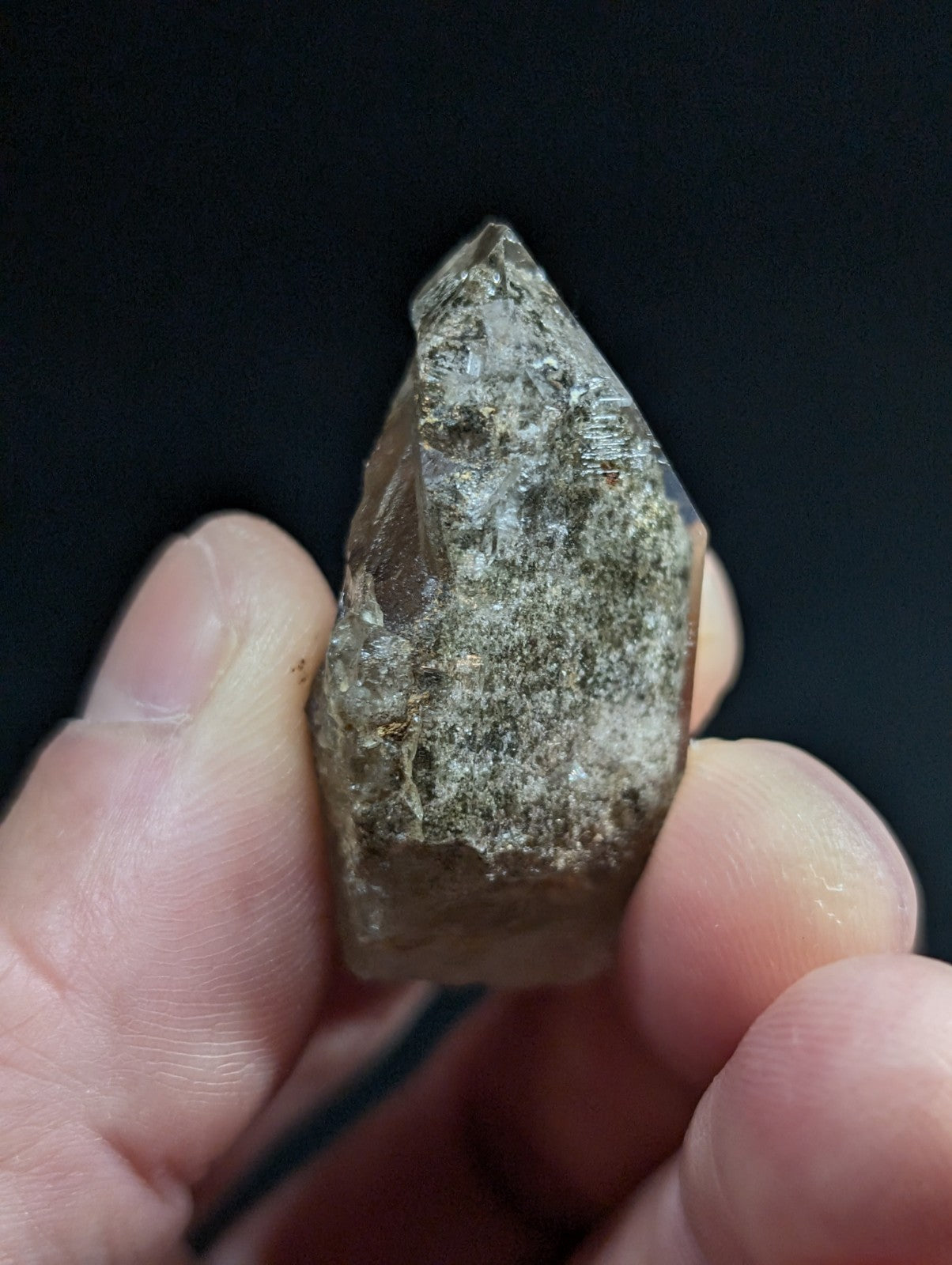 A translucent, pointed quartz crystal with a rough, earthy surface and subtle greenish-gray veining.