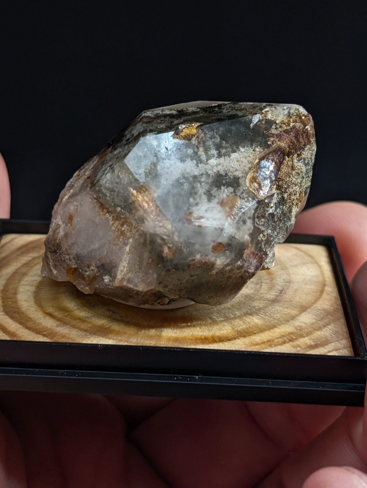 A translucent, grayish-white crystal with golden inclusions and rough, natural facets rests on a wooden display base.