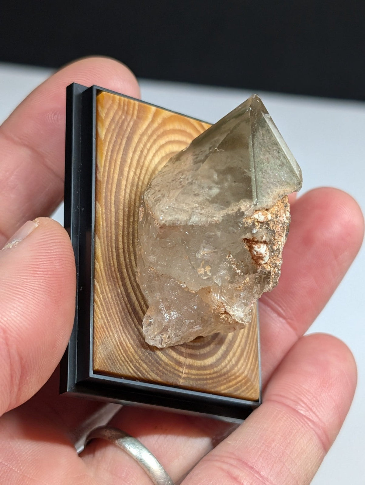 A translucent, pale greenish quartz crystal with a rough, earthy base and sharp geometric facets is mounted in a black-framed wooden display.