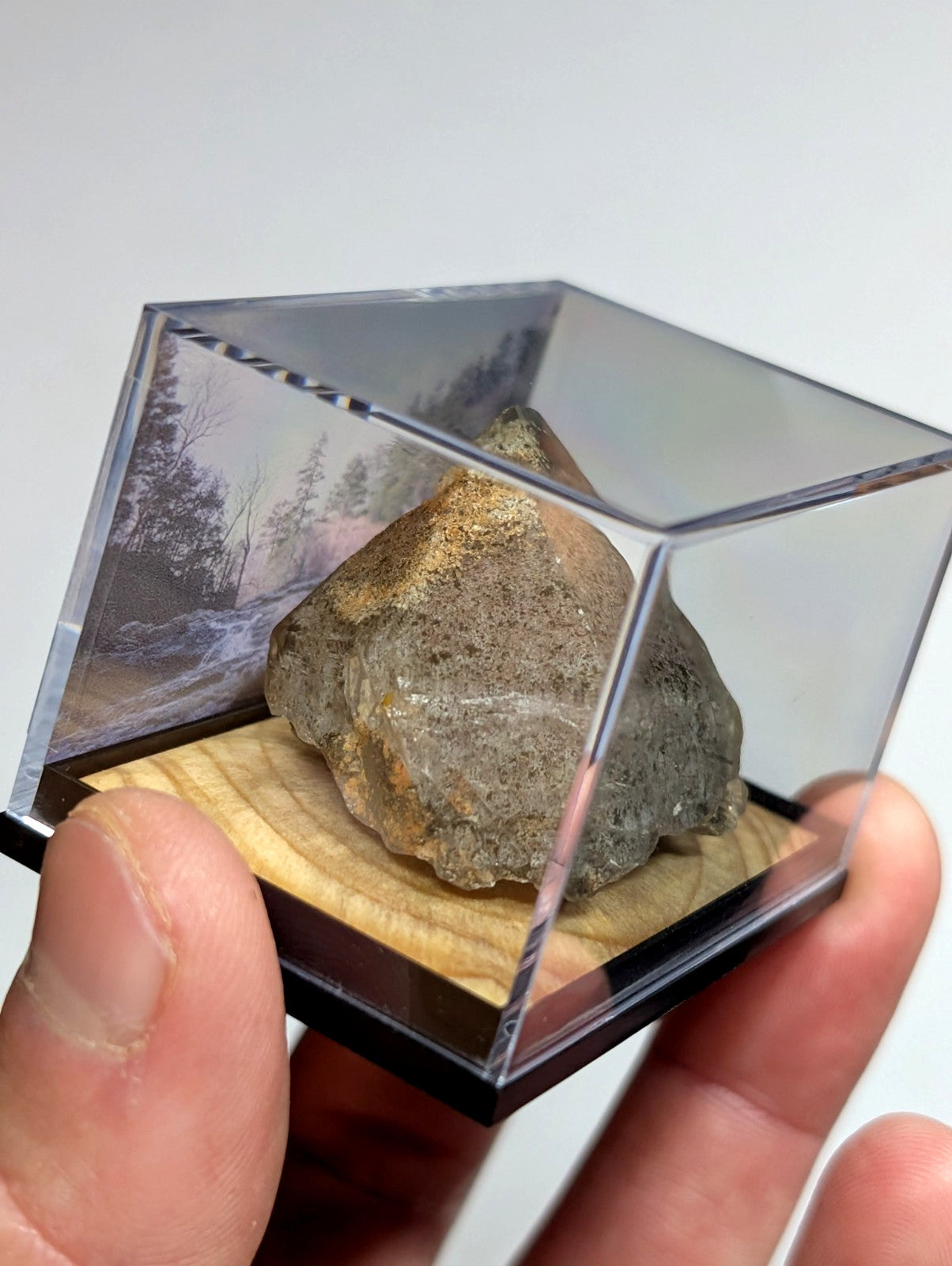 A rugged, grayish-brown rock with a rough texture and subtle orange speckles is displayed inside a clear acrylic cube.