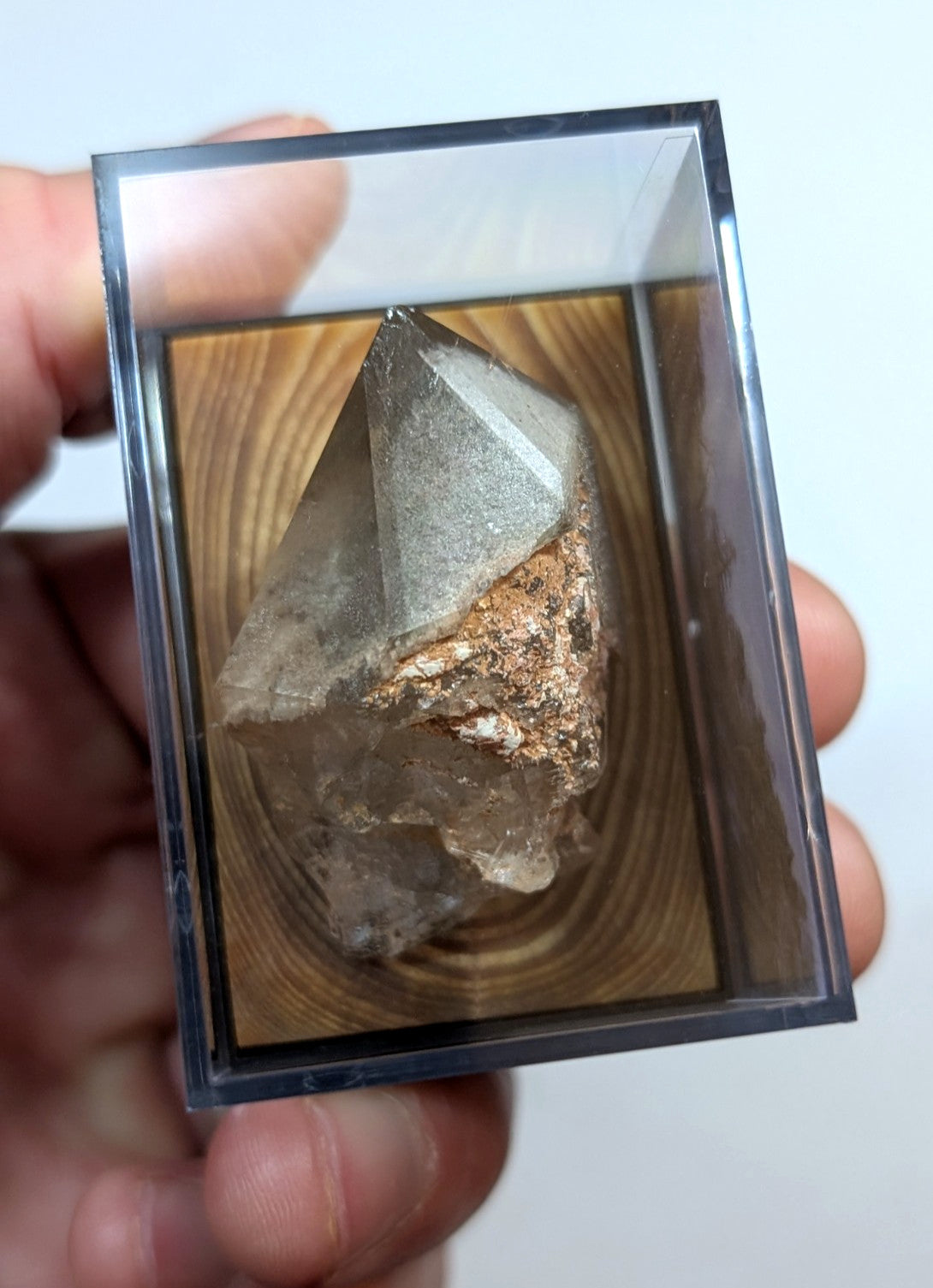 A clear, faceted quartz crystal with a metallic coppery mineral inclusion rests inside a transparent acrylic display case.