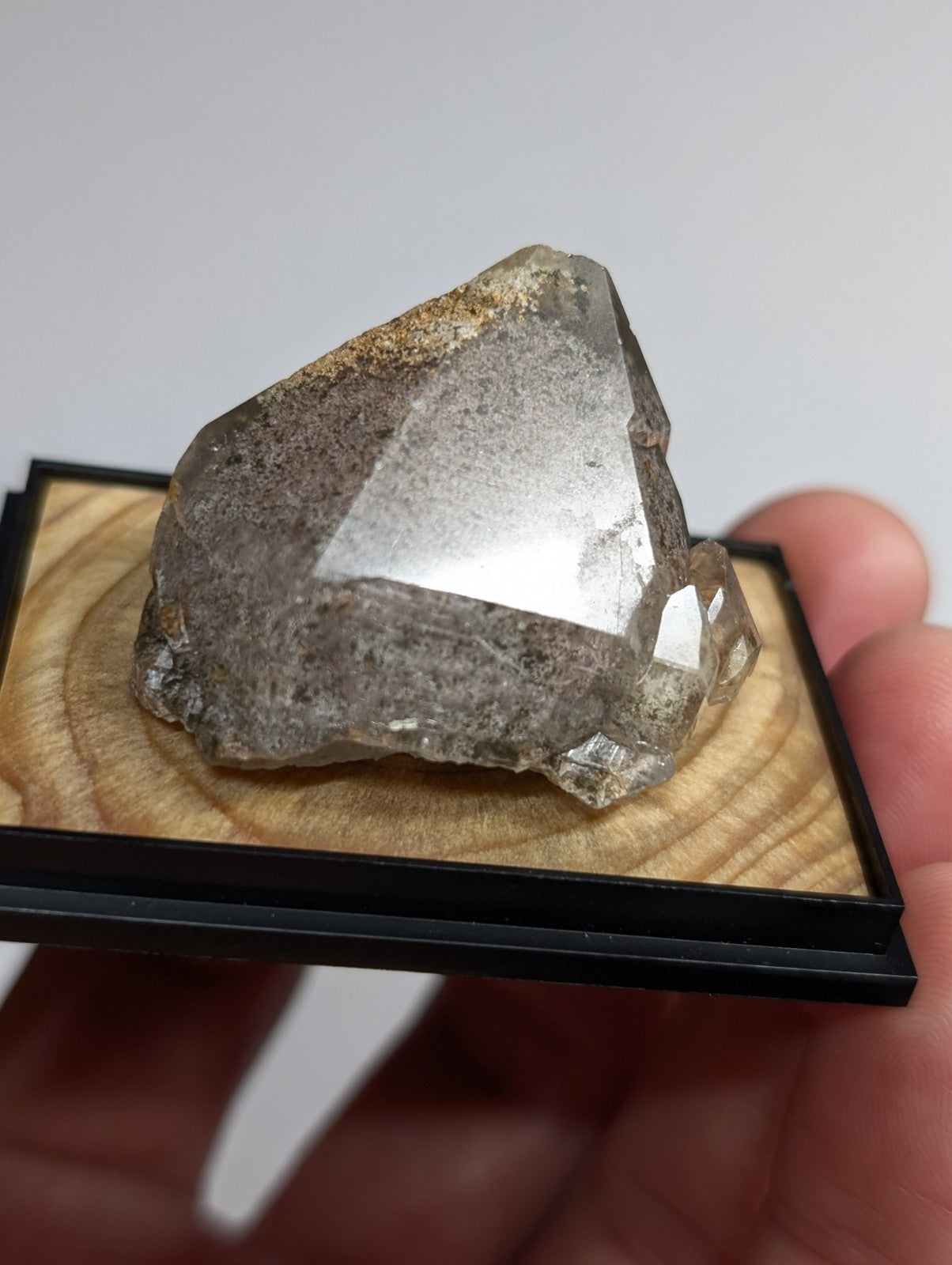 A translucent gray quartz crystal with a rough, faceted surface and subtle brownish inclusions rests on a wooden display base within a black frame.