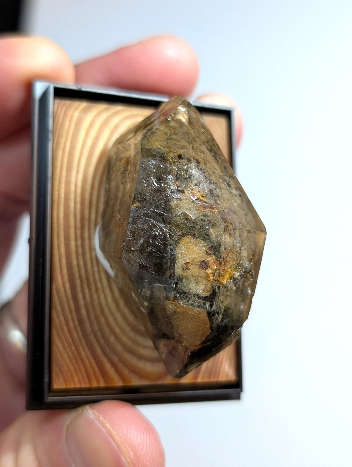 A translucent, faceted crystal with earthy brown and golden inclusions is displayed within a sleek black frame set against a light wood-grain background.