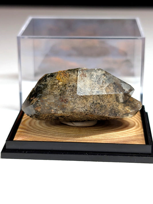 A rugged, translucent quartz crystal with earthy brown and gray veining rests in a clear acrylic display case atop a wooden base.