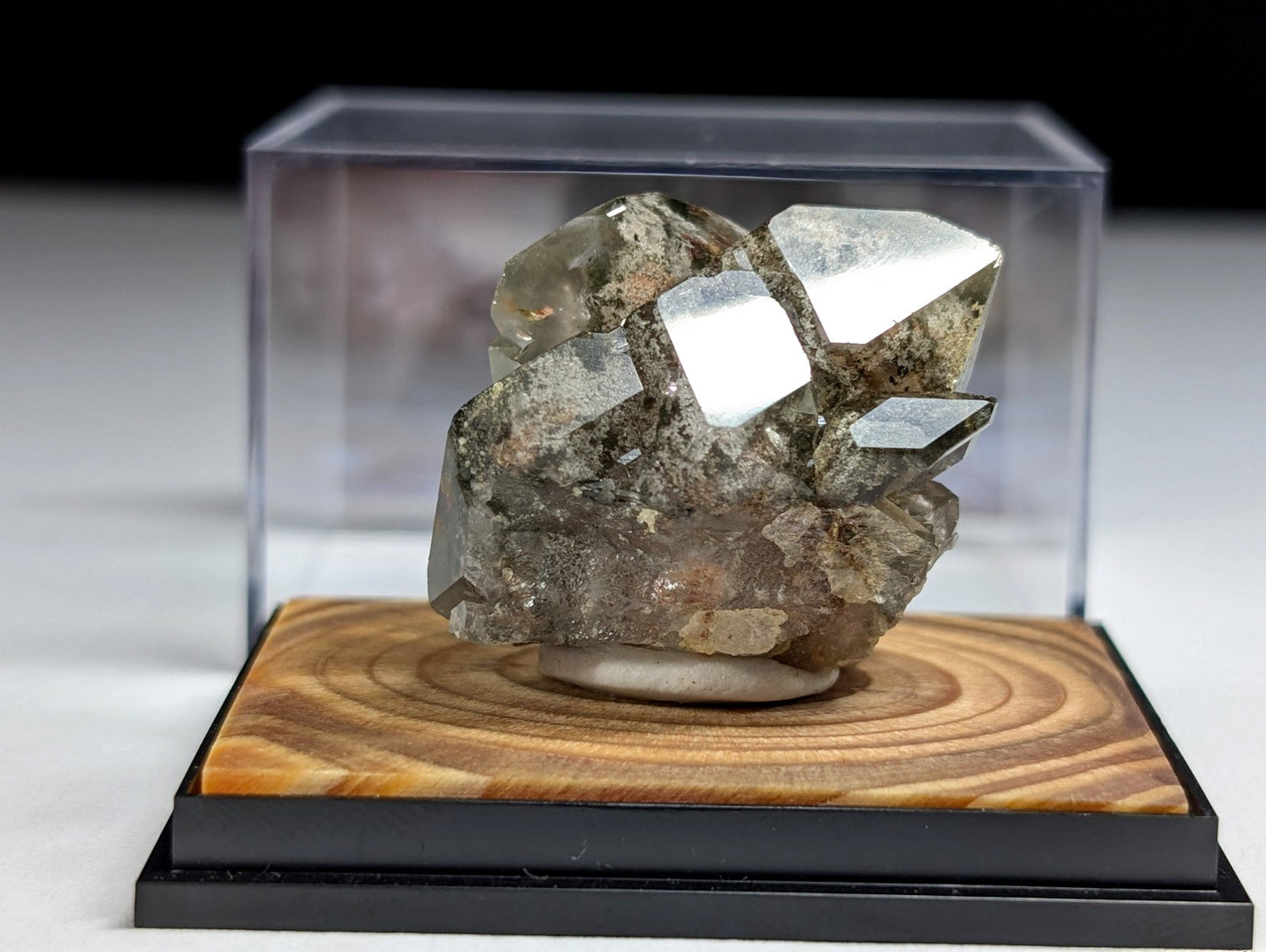 A clear, faceted quartz crystal cluster with a grayish-brown matrix sits elegantly displayed in a transparent acrylic case atop a polished wooden base.