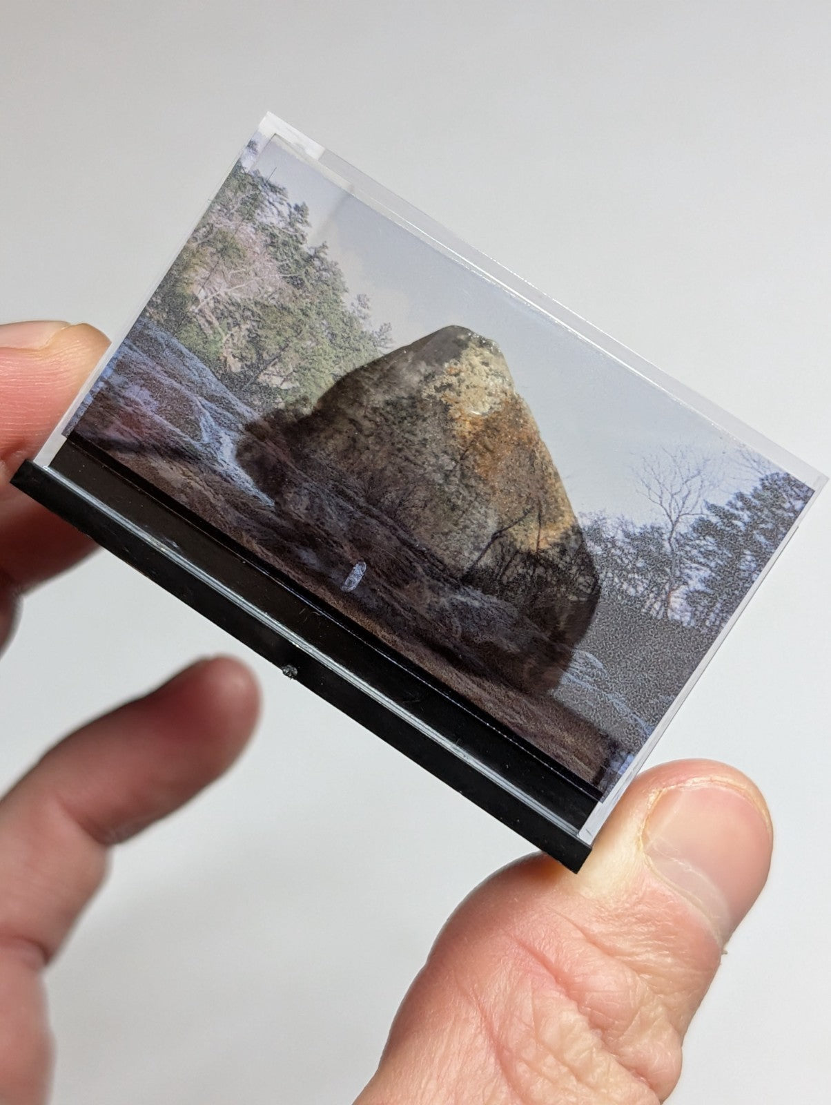 A rectangular, transparent photo frame with a black base holding an image of a large mossy rock in a forest setting.