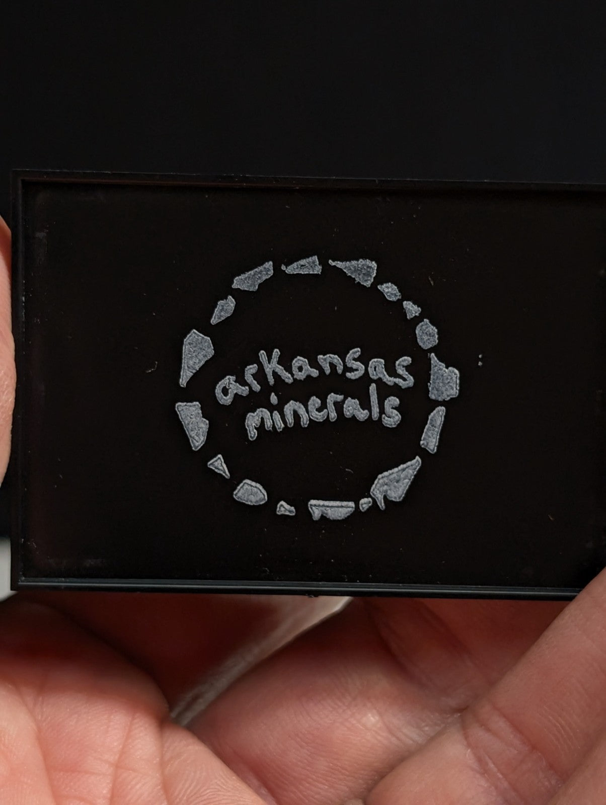 Black rectangular compact with silver engraved ’arkansas minerals’ logo surrounded by a circle of stylized mineral shapes.