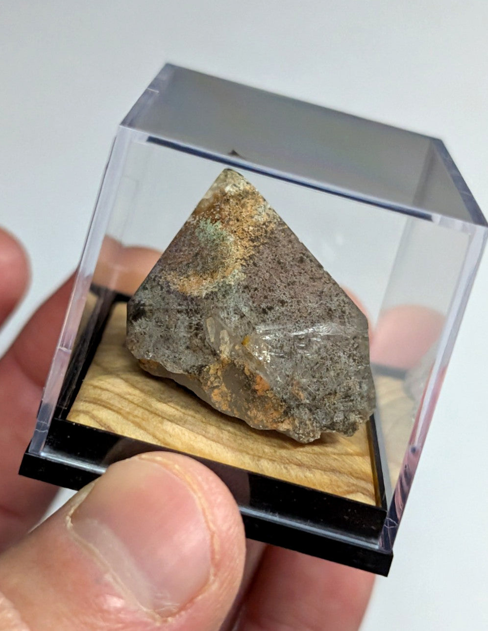 A triangular, rough-textured mineral specimen with mottled gray and brown hues is displayed in a clear acrylic cube.