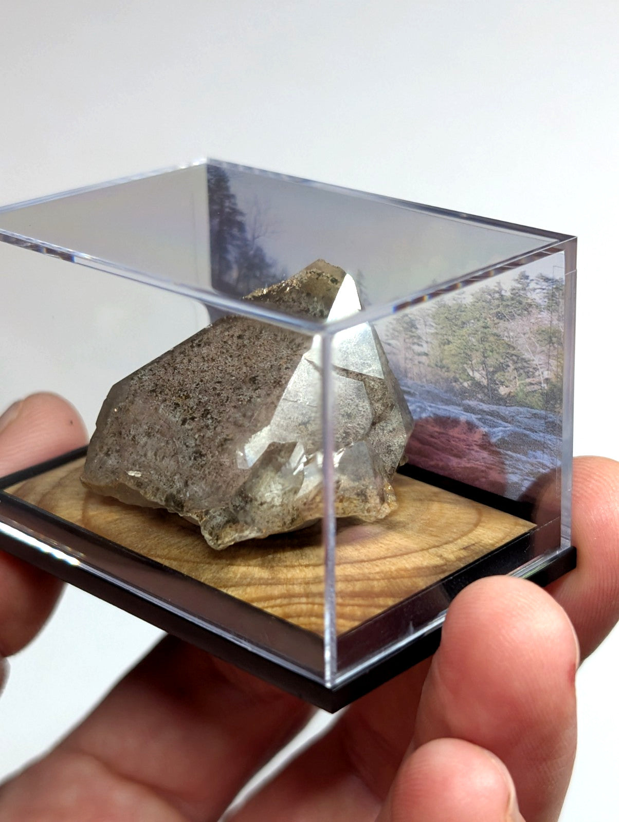 A clear acrylic display case holding a rough, translucent crystal with earthy brown and gray tones resting on a wooden base.