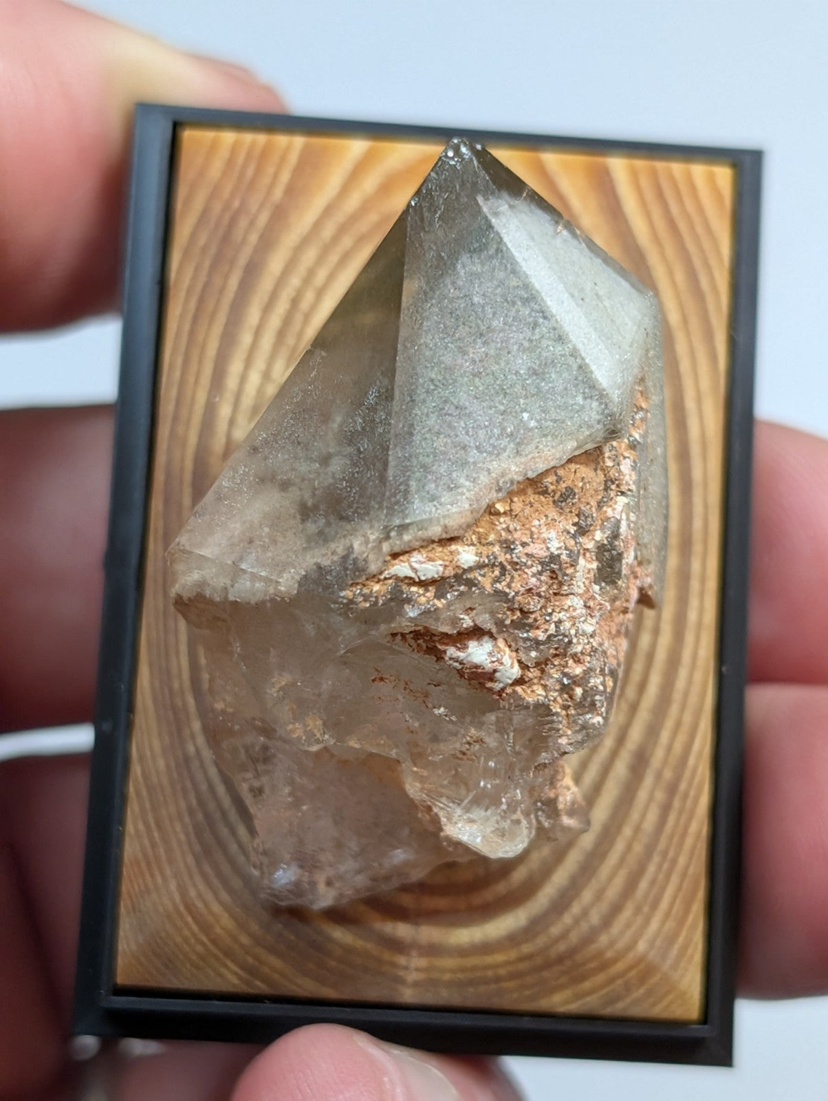 A clear, faceted quartz crystal with a coppery mineral inclusion is mounted in a black-framed wooden display.