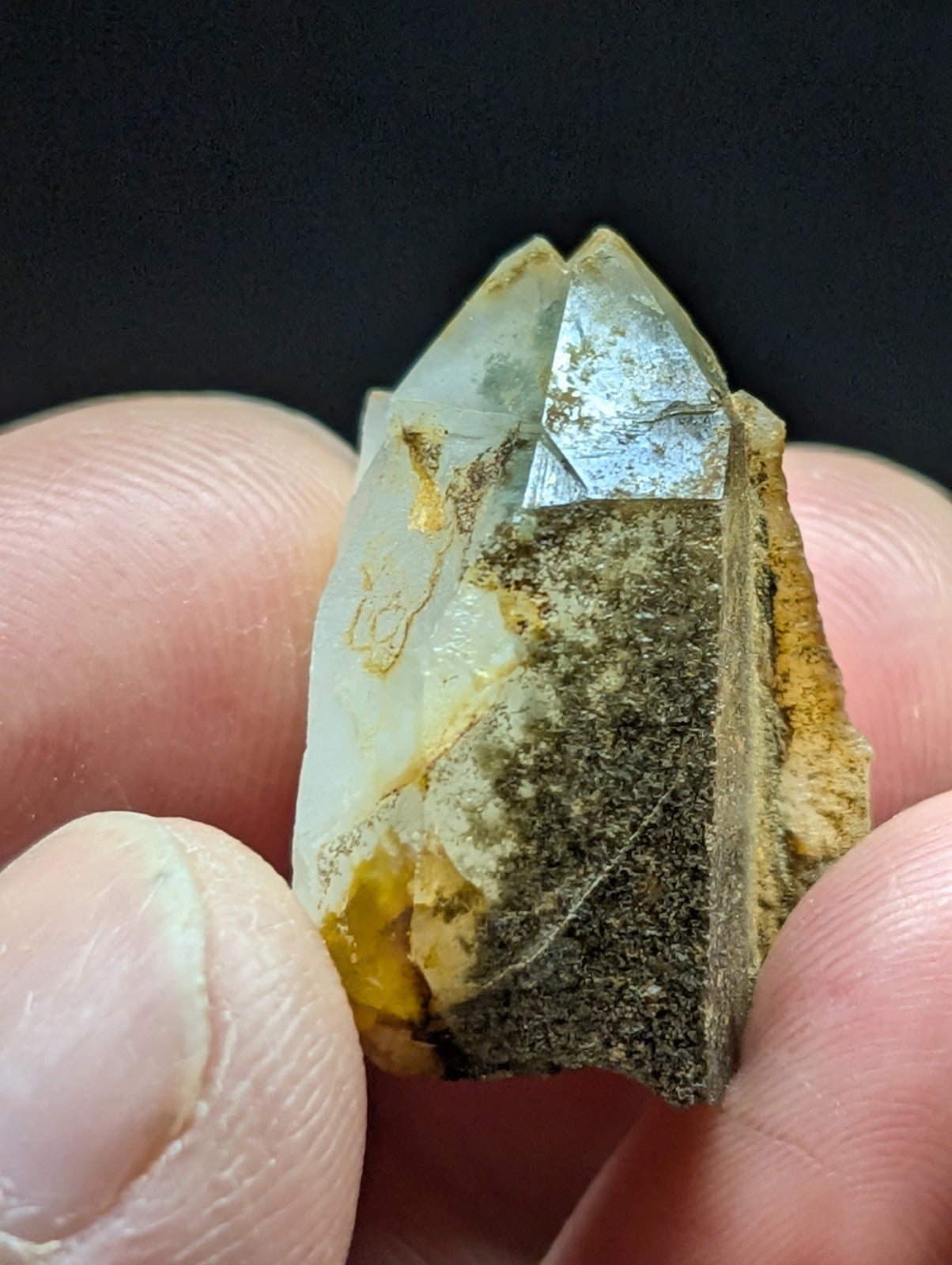A translucent quartz crystal with a pointed tip, featuring natural inclusions of yellow and dark green minerals against its clear base.