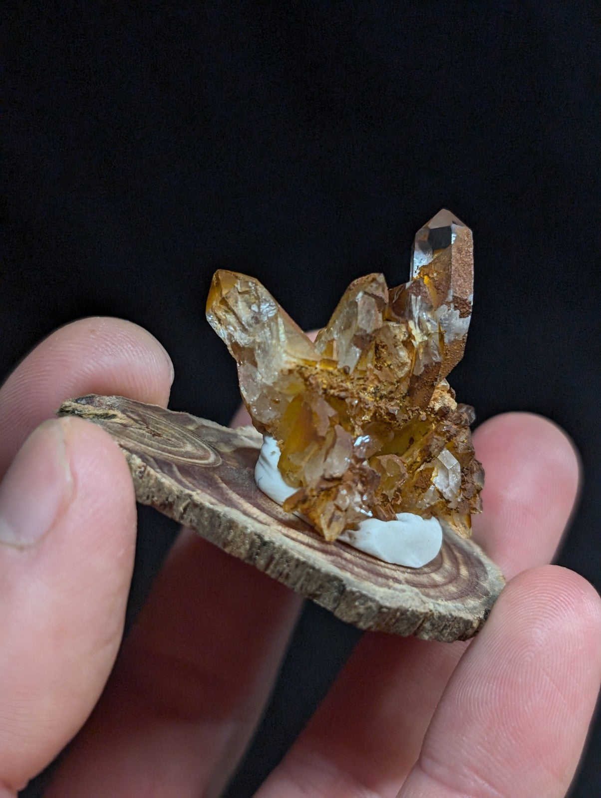 A golden amber crystal cluster with translucent facets and earthy brown matrix, displayed on a rustic wooden slice.