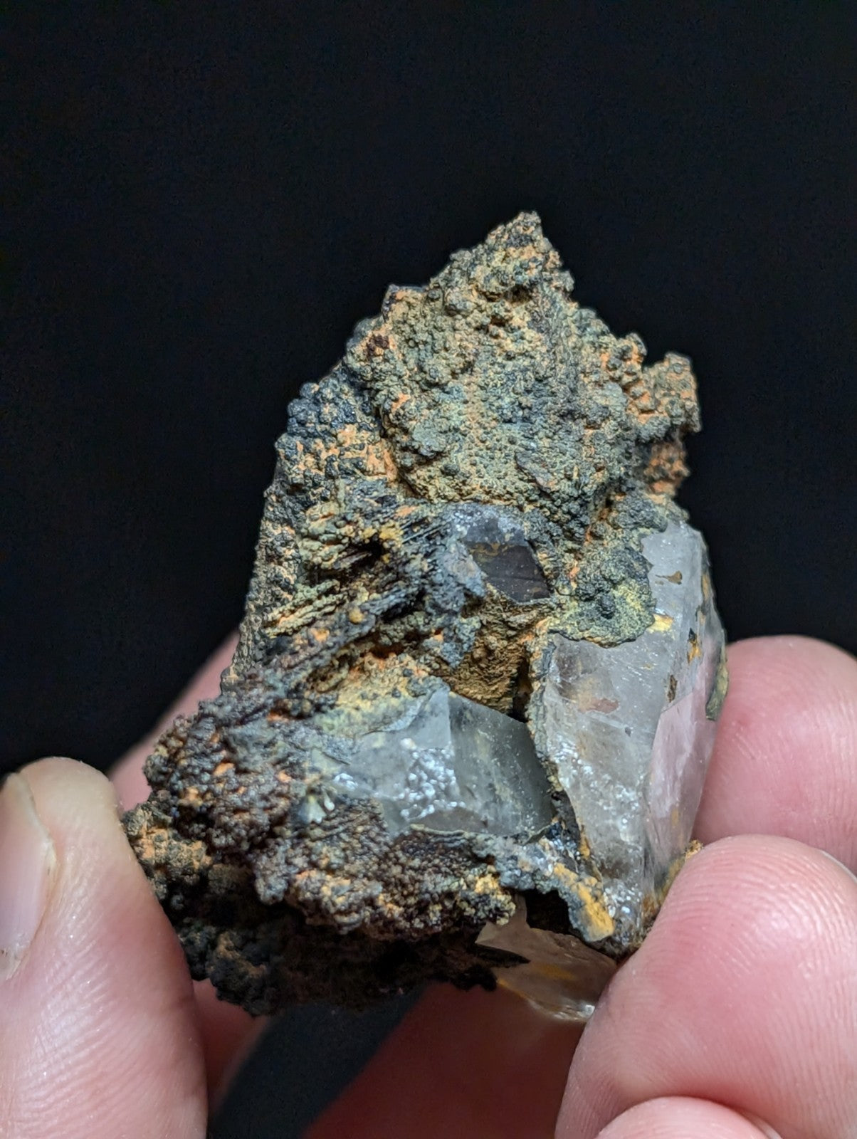 A translucent quartz crystal embedded in a rugged, earthy matrix of dark green and brown mineral formations.