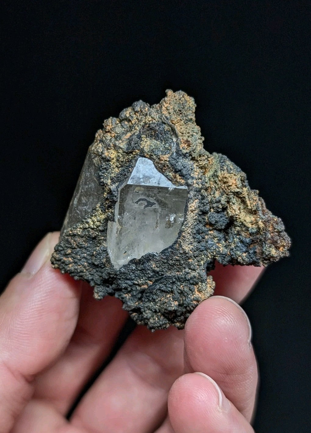 A clear, faceted quartz crystal embedded in a rough, dark gray and brown mineral matrix.