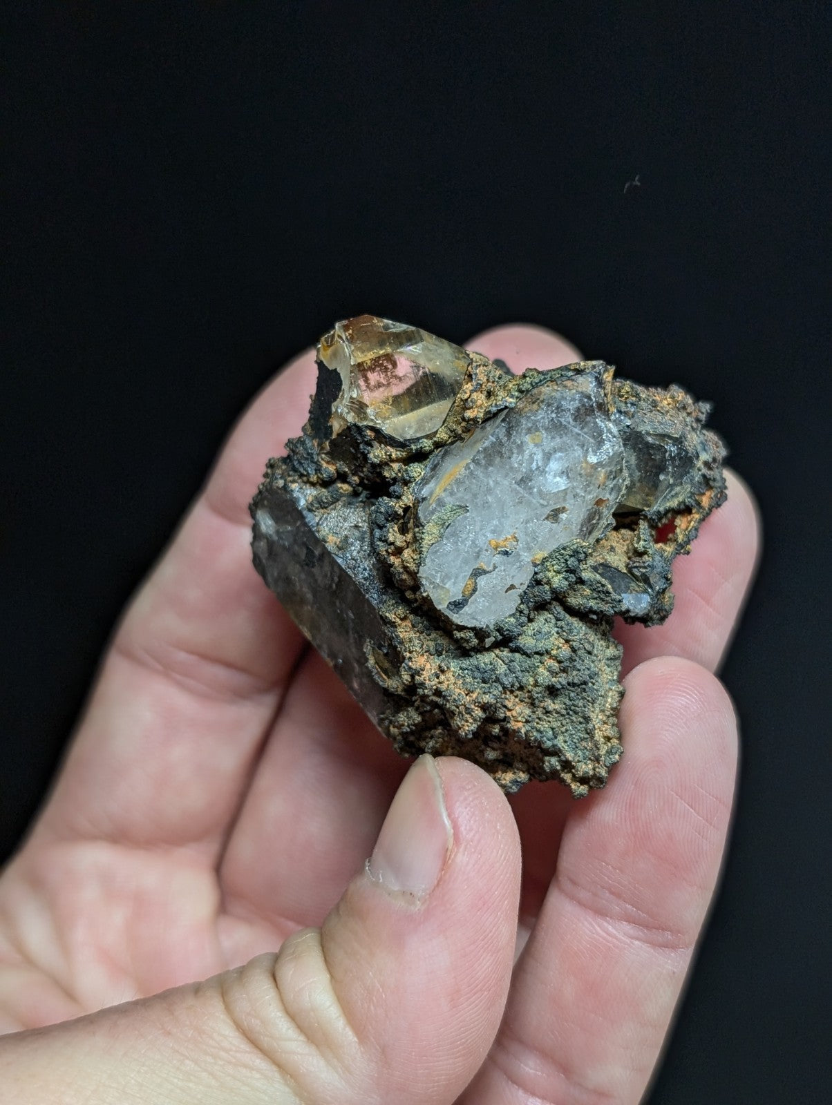 A hand holds a rough, dark mineral specimen featuring translucent quartz crystals with golden-brown and reddish hues embedded in its rocky matrix.