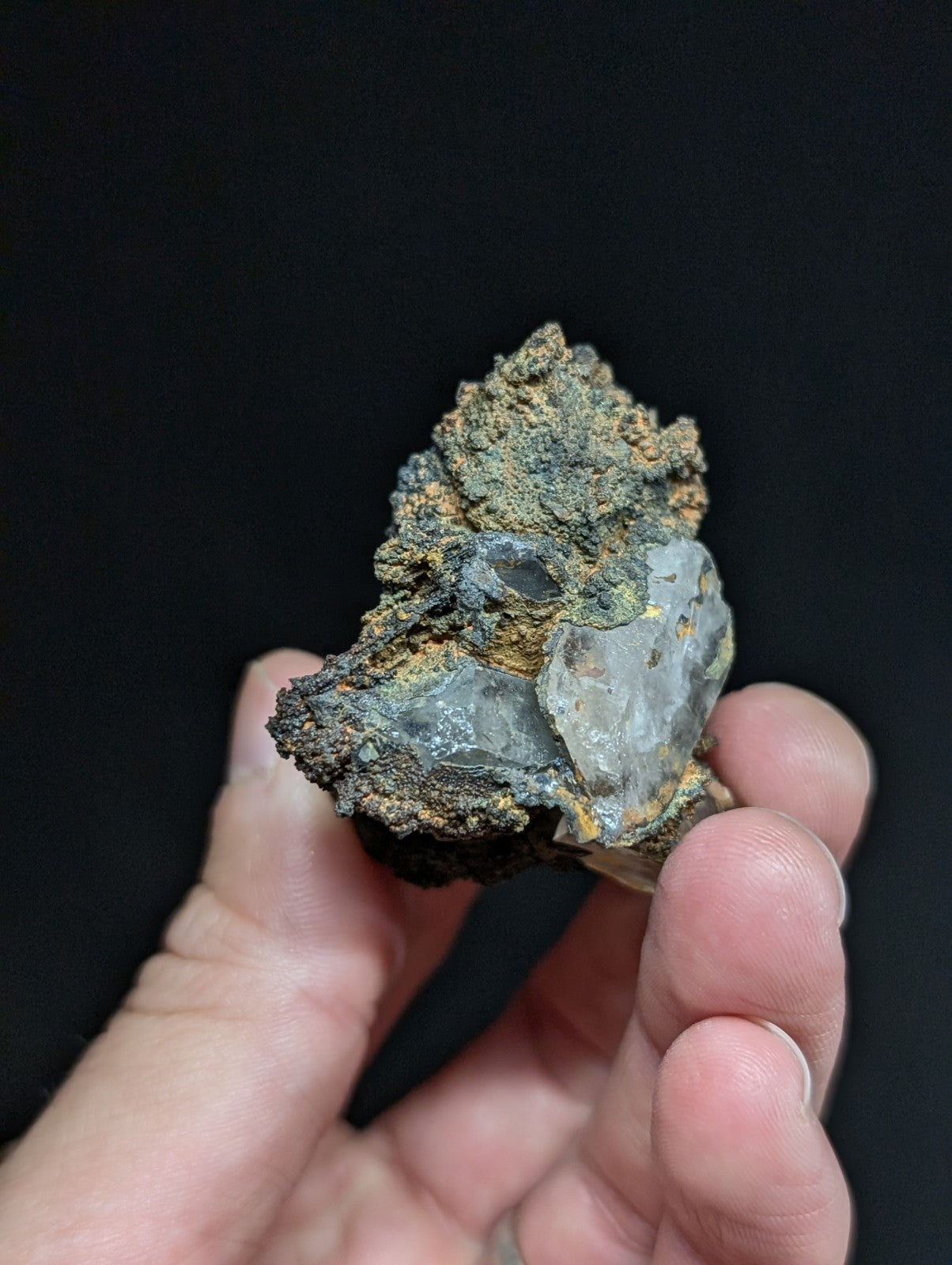 A hand holds a rough, irregularly shaped mineral specimen featuring translucent gray quartz crystals embedded in dark, earthy matrix material with patches of orange and green oxidation.