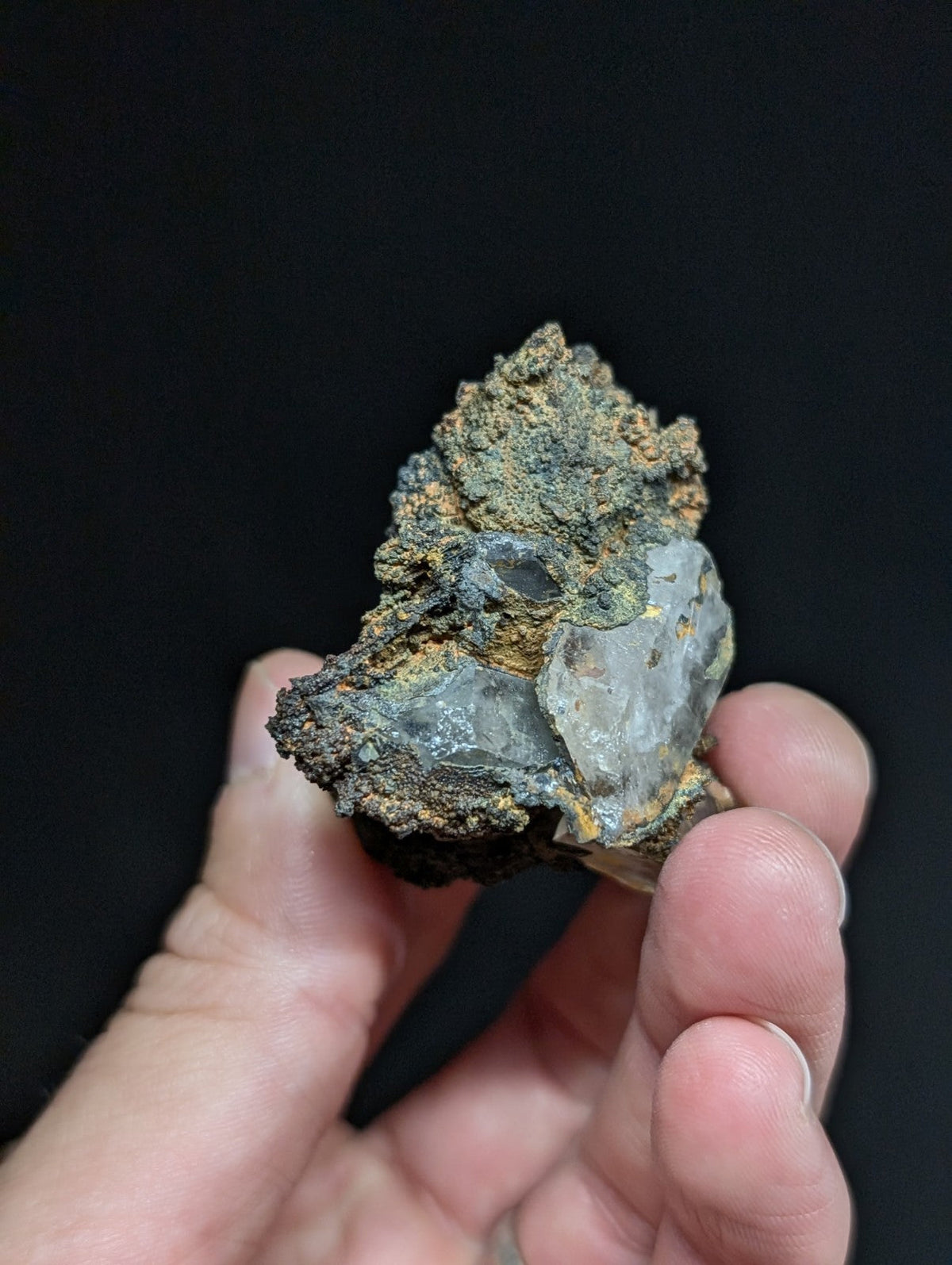 A hand holds a rough, irregularly shaped mineral specimen featuring translucent gray quartz crystals embedded in dark, earthy matrix material with patches of orange and green oxidation.
