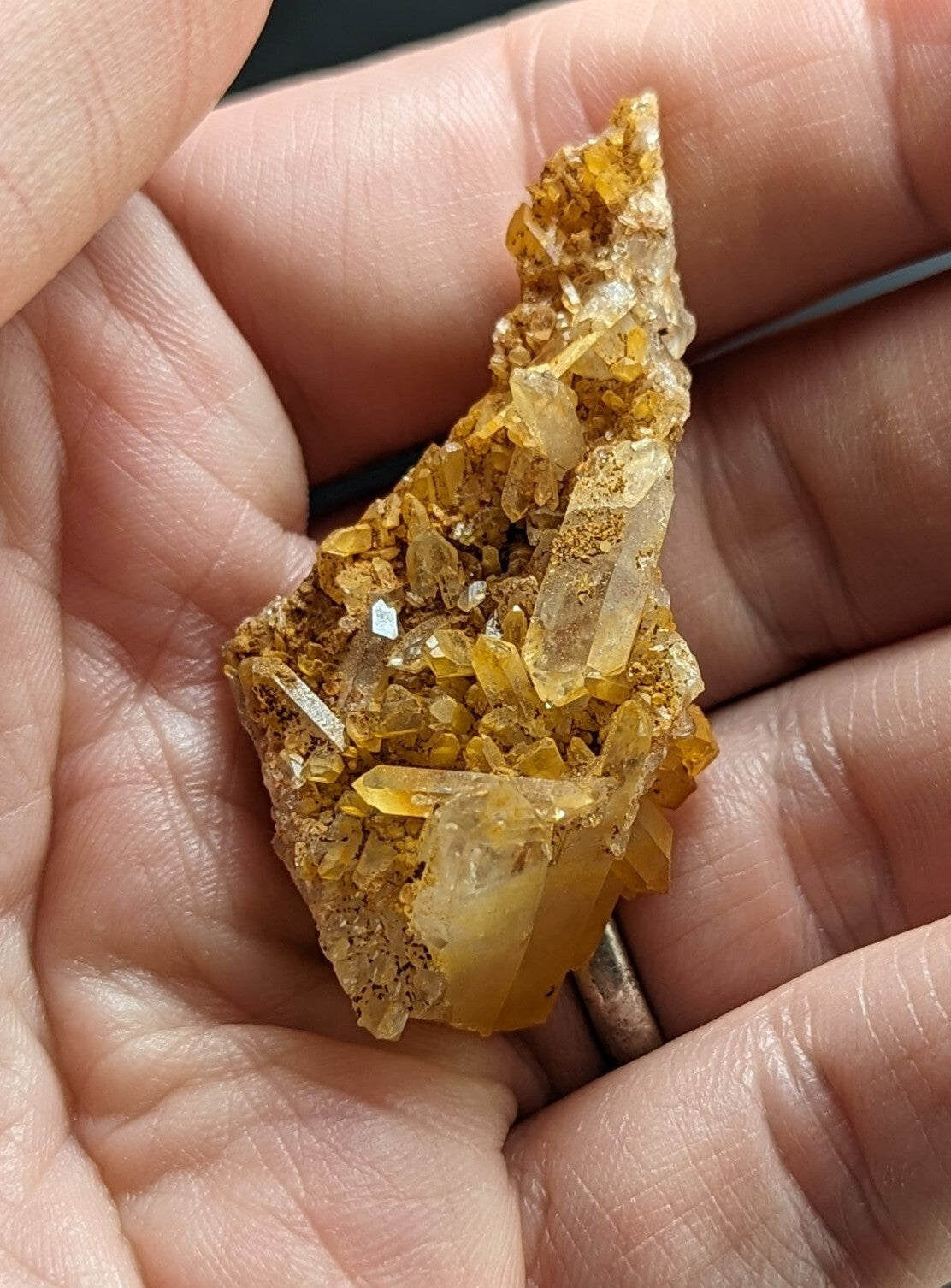 A hand holds a cluster of translucent golden-yellow crystals with clear, faceted points emerging from a rougher, earthy matrix.