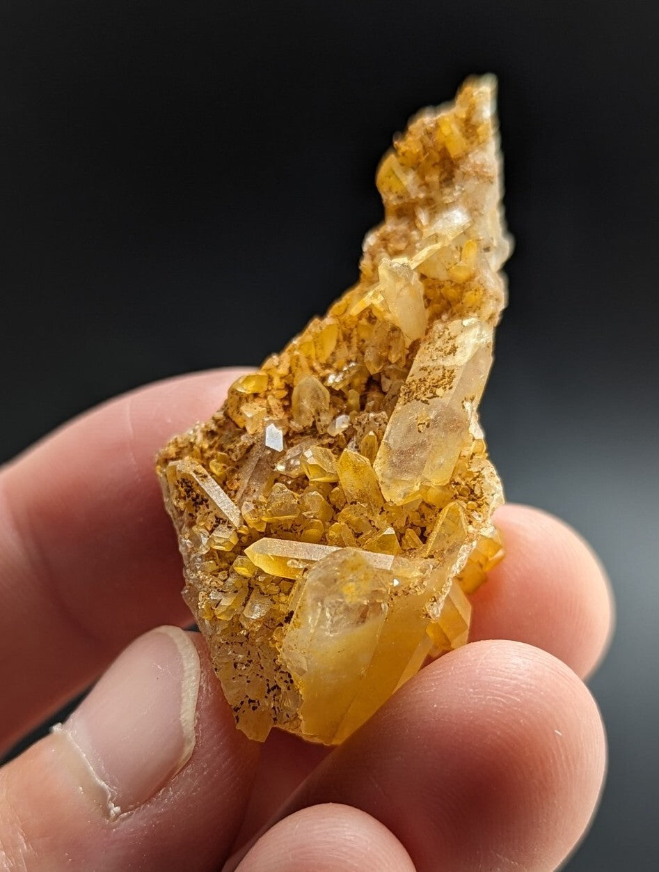 A hand holds a translucent golden-yellow crystal cluster with sharp, faceted points and embedded mineral inclusions.
