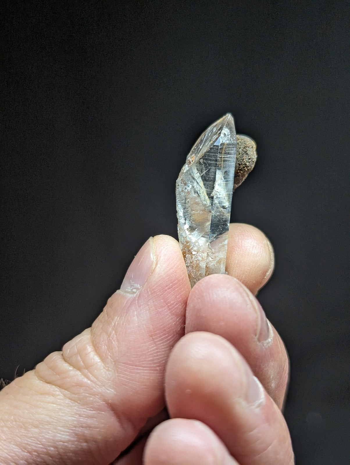 A clear quartz crystal with a pointed tip and visible internal striations, held gently between fingers against a dark background.