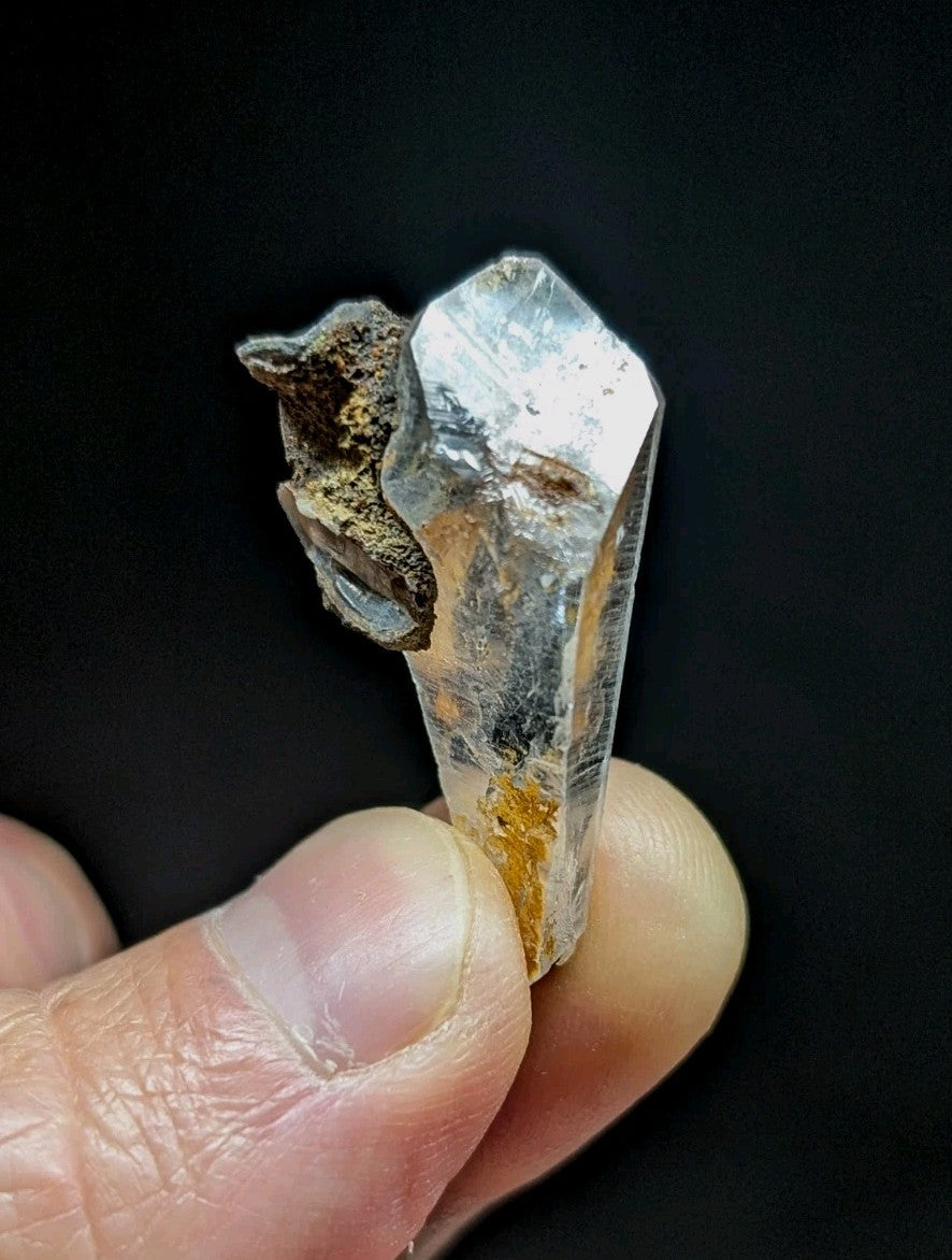 A translucent quartz crystal with a pointed tip, featuring natural inclusions and earthy brown mineral deposits along its base.