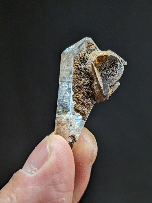 A clear quartz crystal with a rough, earthy brown matrix growing from its top.