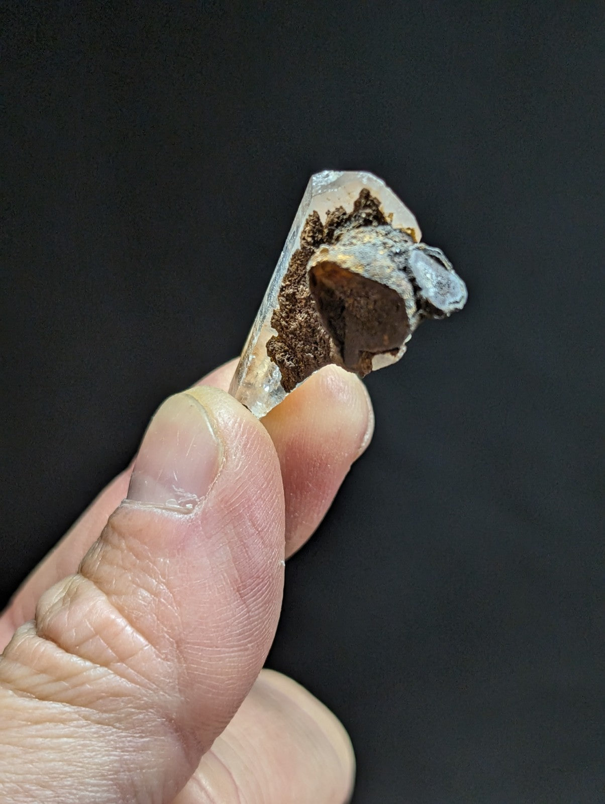 A translucent quartz crystal with a rough, earthy brown and gray mineral inclusion at its base.