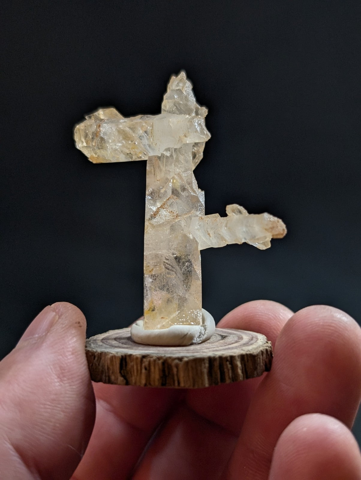 A translucent, pale yellow crystal formation shaped like a stylized cross or t-shape stands on a circular wooden base.