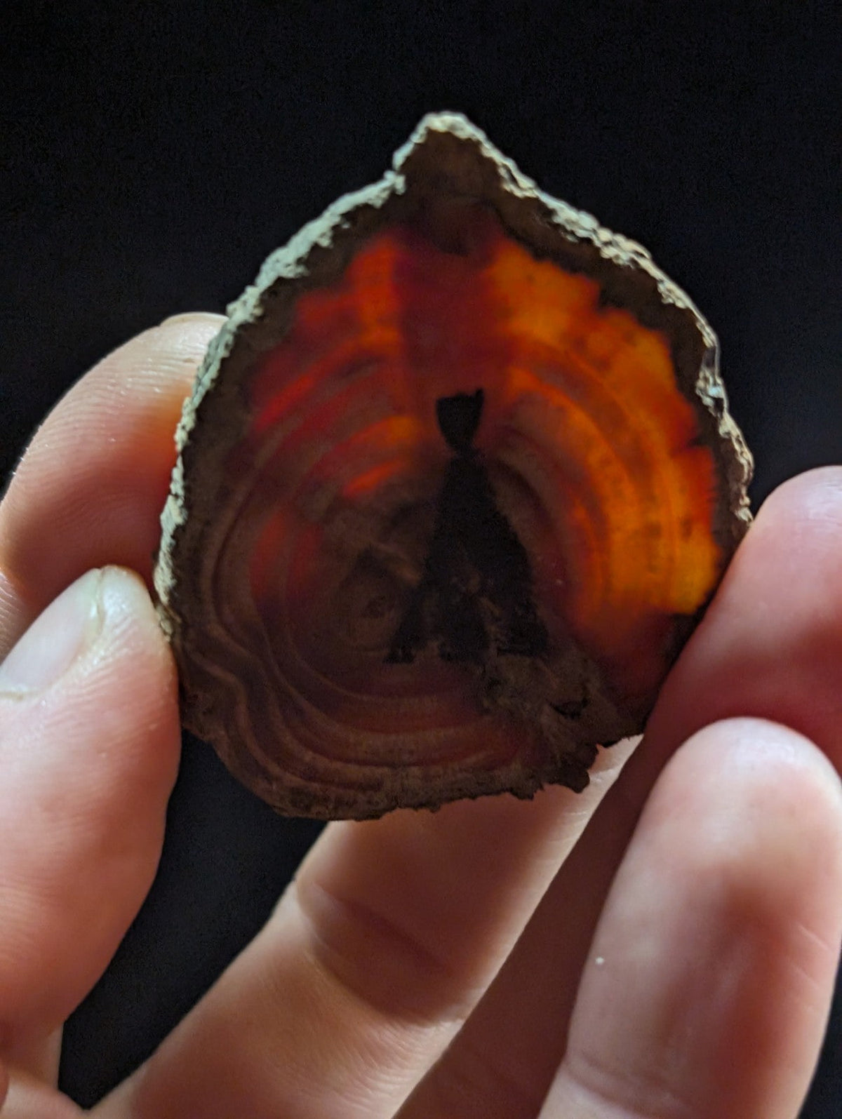 A hand holds a slice of orange and red agate with concentric rings, featuring a dark silhouette of a figure at its center.