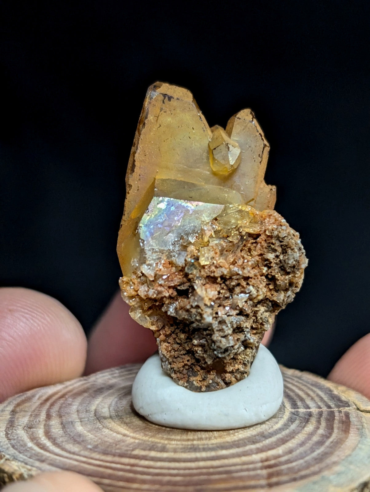 A translucent golden-yellow crystal formation with a shimmering, iridescent opal-like inclusion nestled atop a rough-textured brownish mineral base.
