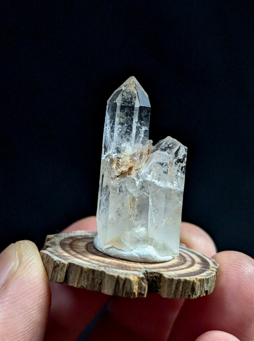 A clear quartz crystal cluster with a pointed, faceted top and natural imperfections sits atop a rustic wooden slice.