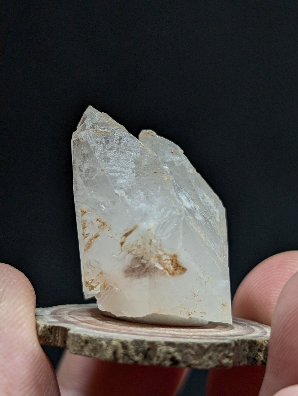 A translucent, clear quartz crystal with subtle brown inclusions and a pointed tip, displayed on a rustic wooden slice.