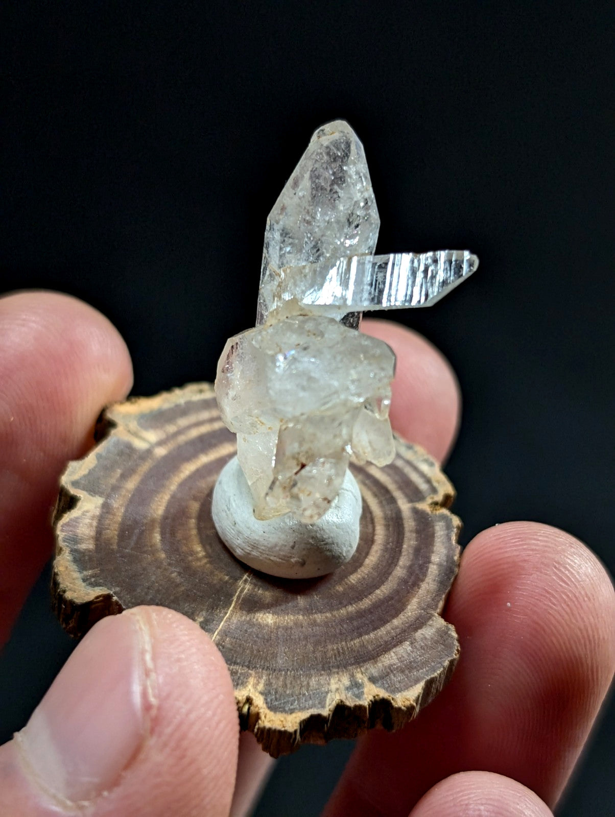 A clear quartz crystal cluster with a pointed, faceted top and layered base, mounted on a small white pedestal atop a circular wooden slice.