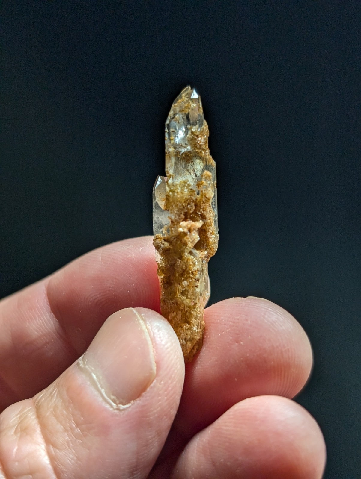 A translucent quartz crystal with a pointed tip and earthy brown matrix clinging to its surface, held gently between fingers.