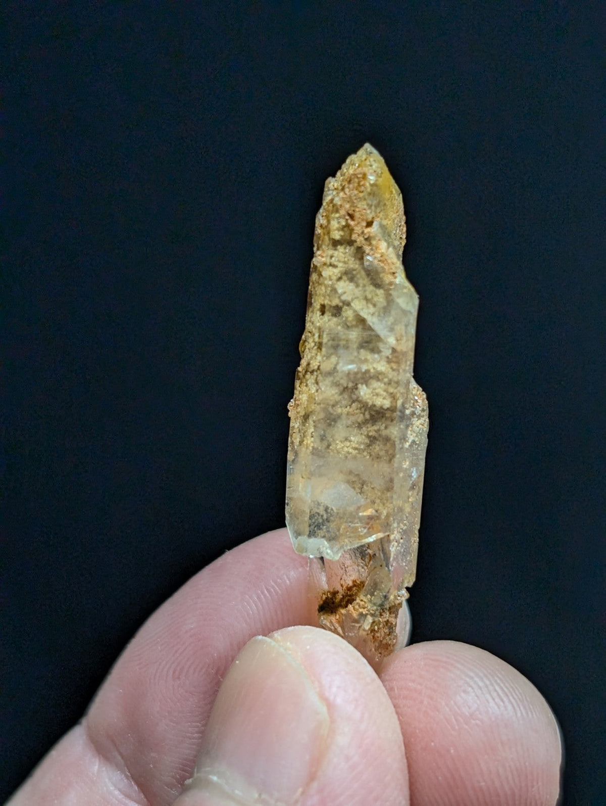 A translucent yellowish quartz crystal with a pointed tip and natural mineral inclusions, held gently between fingers against a dark background.