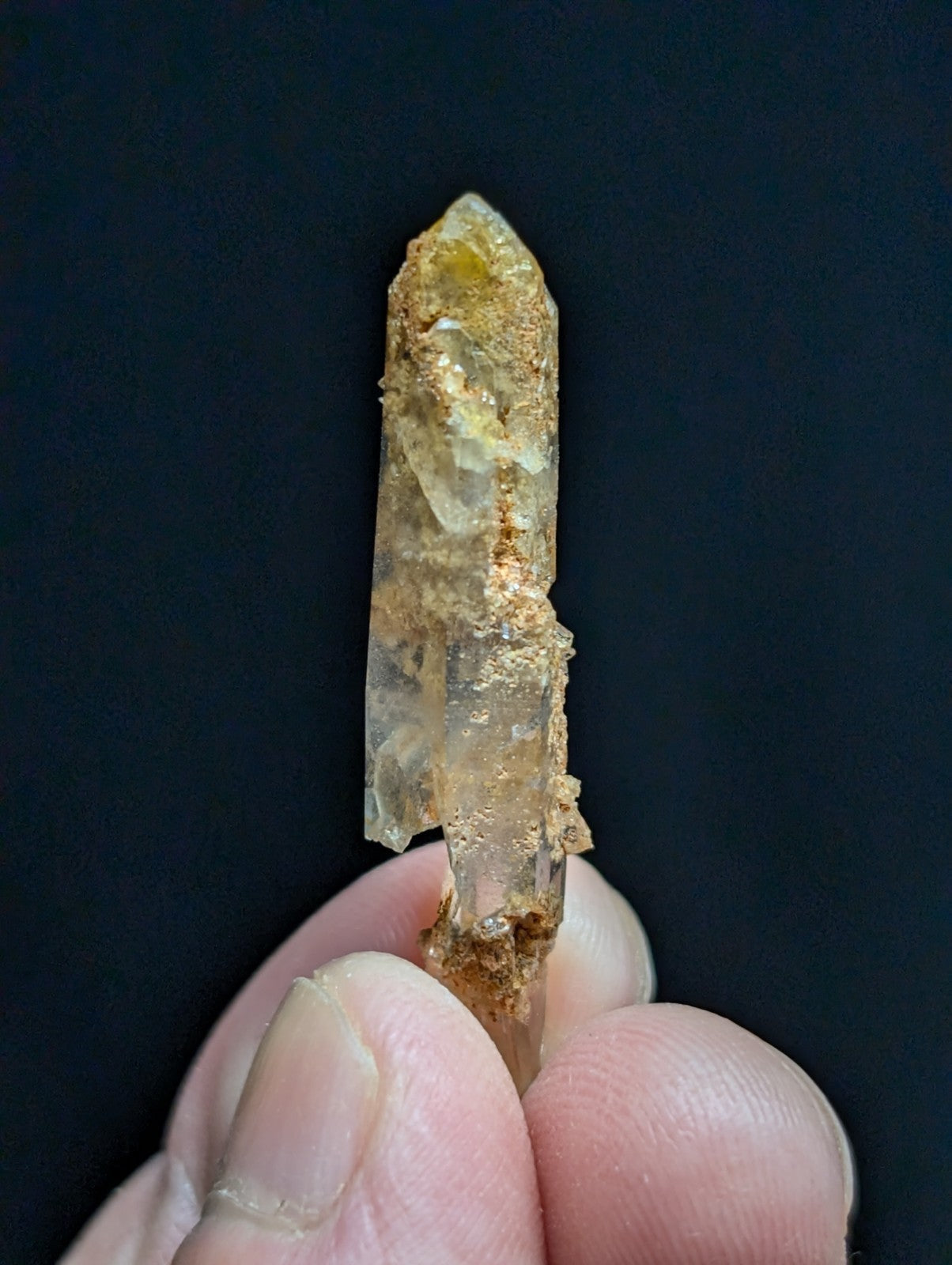 A translucent yellowish quartz crystal with a pointed tip and earthy brown mineral residue clinging to its surface, held gently between fingers.