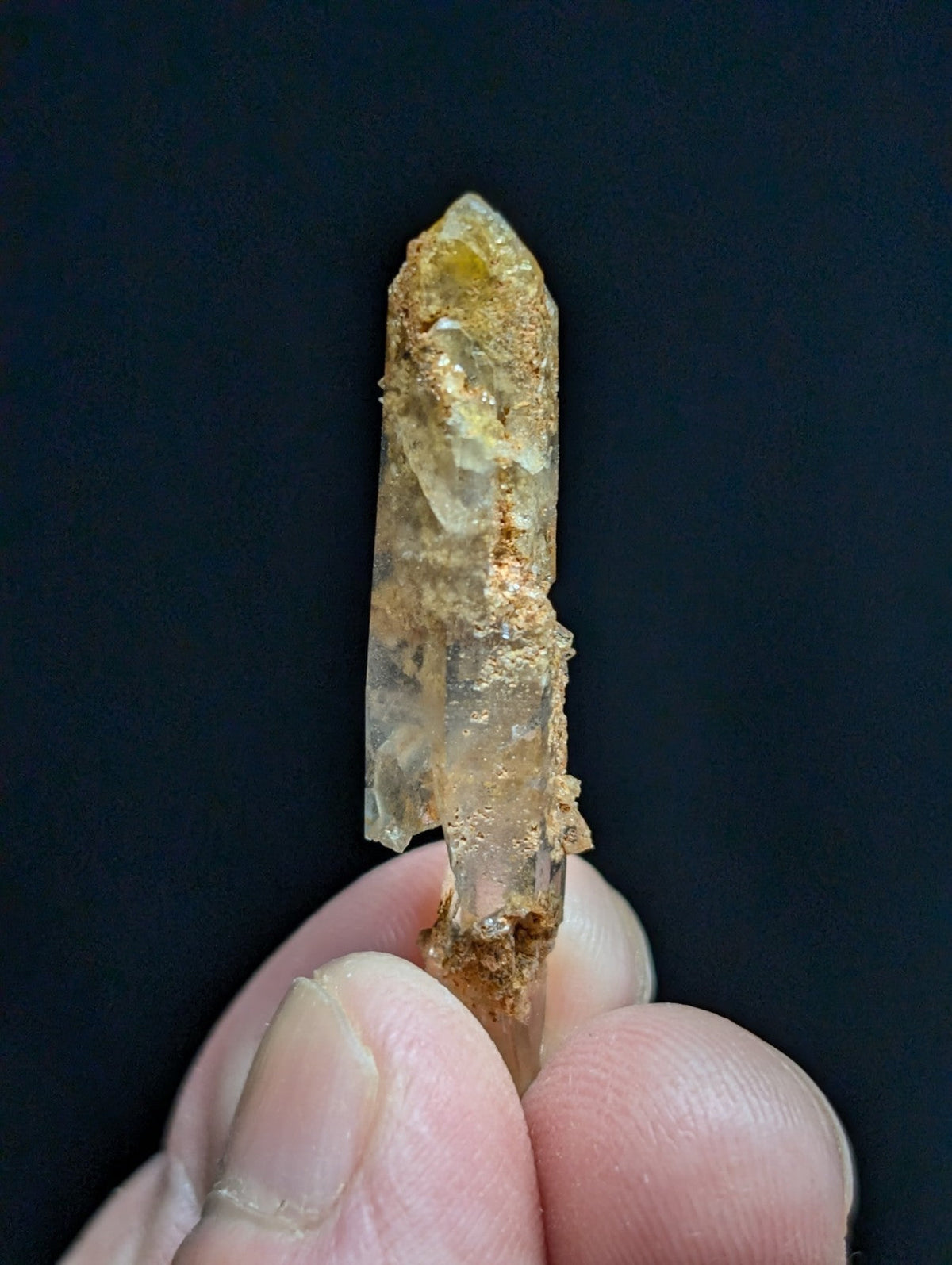 A translucent yellowish quartz crystal with a pointed tip and earthy brown mineral residue clinging to its surface, held gently between fingers.