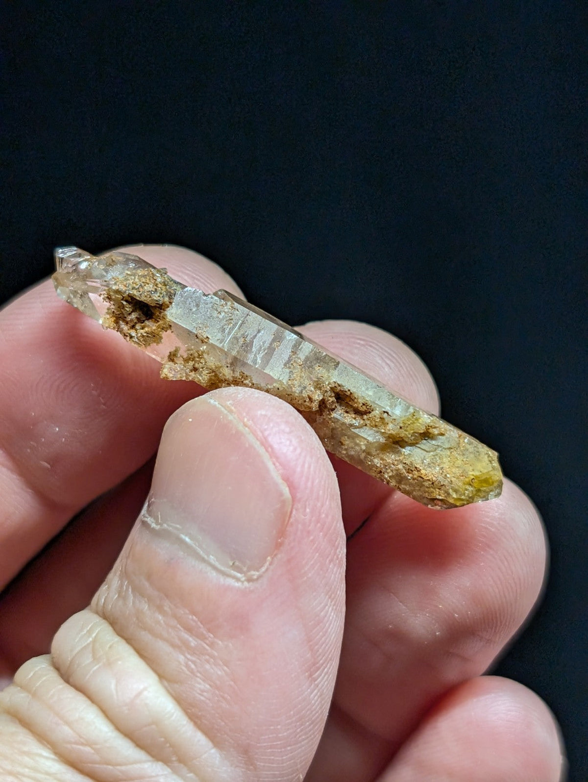 A translucent quartz crystal with natural yellowish-brown mineral inclusions and striations, held gently between fingers.