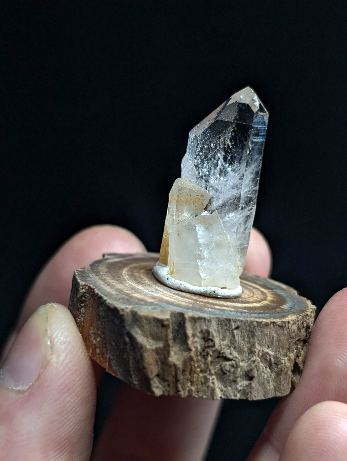 A clear quartz crystal with a pointed tip and subtle internal veining, mounted on a rustic wooden slice base.