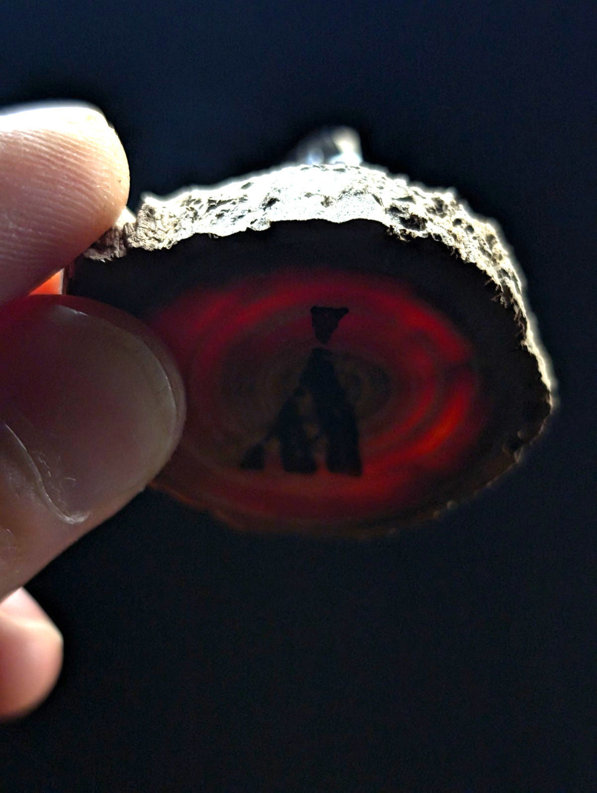A hand holds a slice of red agate with a rough, natural edge and an intricate black silhouette design etched into its center.
