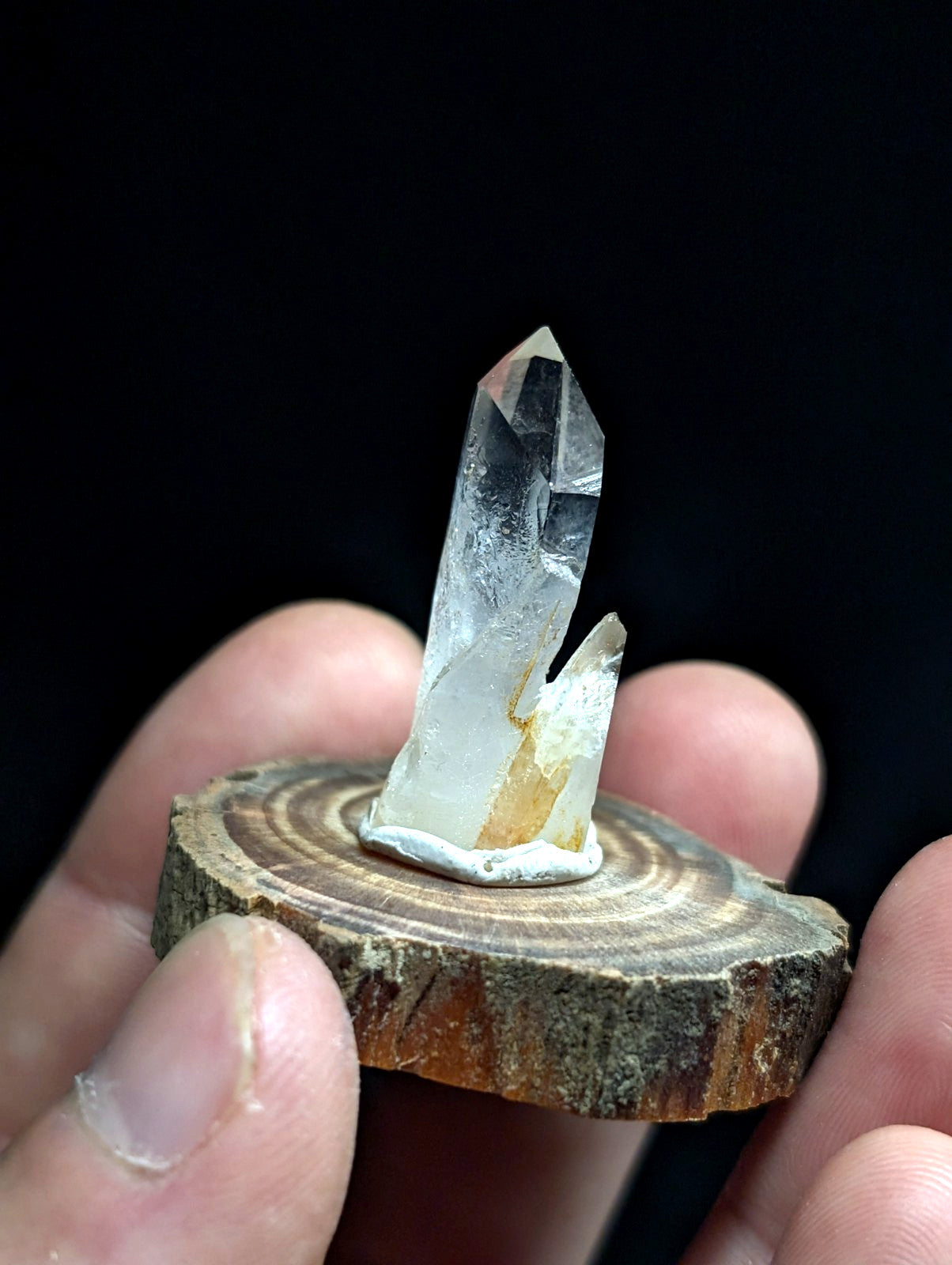 A clear quartz crystal with a pointed tip and subtle internal inclusions, mounted on a rustic wooden slice.