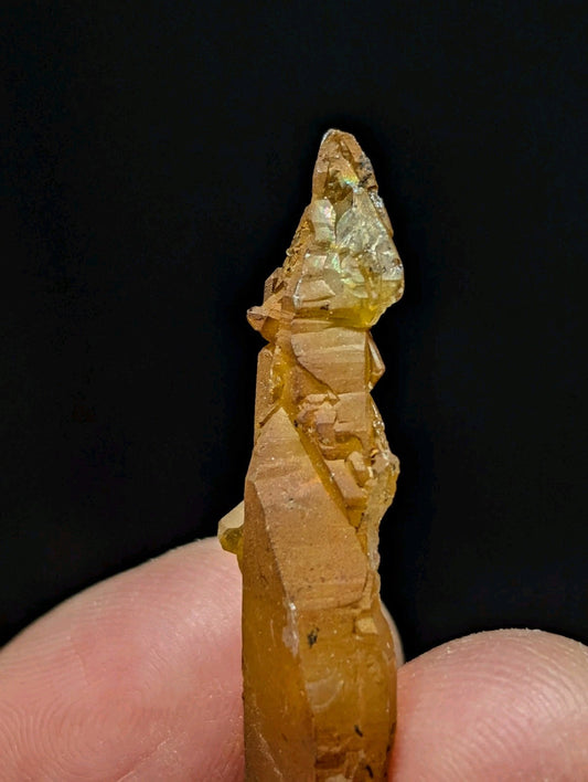 A translucent amber-colored crystal with a pointed, faceted top and layered striations along its length.