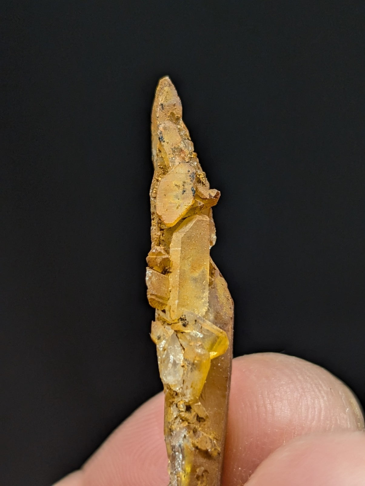 A translucent yellow crystal with a pointed tip and embedded facets, held between fingers against a dark background.