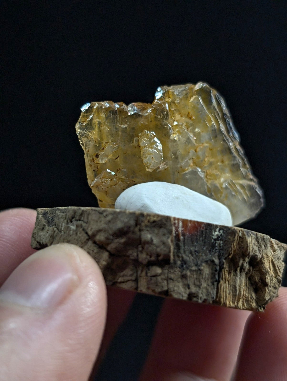 A translucent amber-colored crystal with a rough, faceted surface rests atop a smooth white stone, both displayed on a rustic wooden slice held in hand.