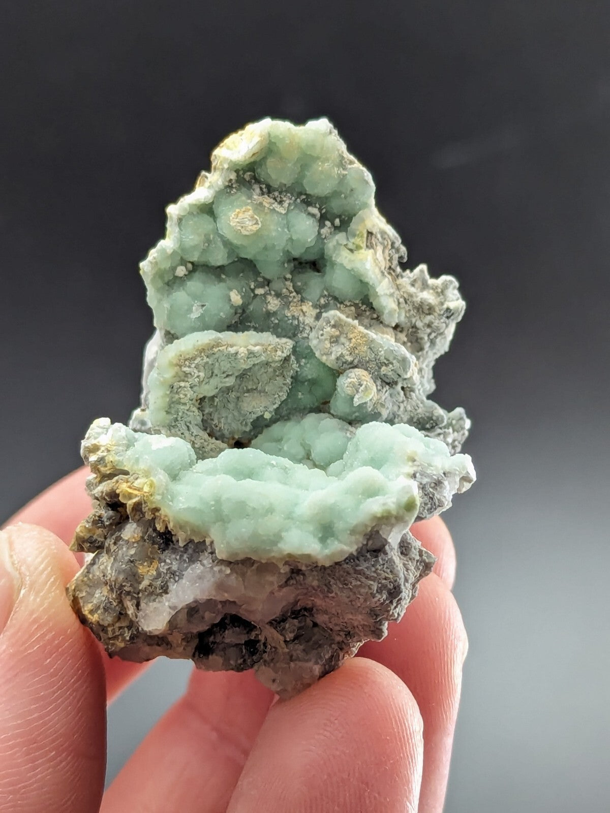 A hand holds a natural mineral specimen featuring light green, clustered crystals growing atop a darker gray rock matrix.