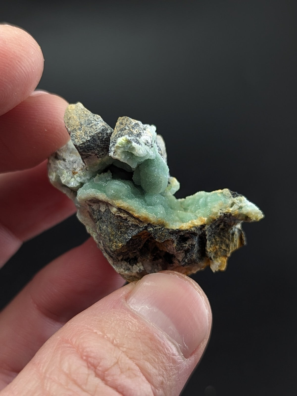 A hand holds a natural mineral specimen featuring translucent green crystals growing from a dark, rough-textured matrix.
