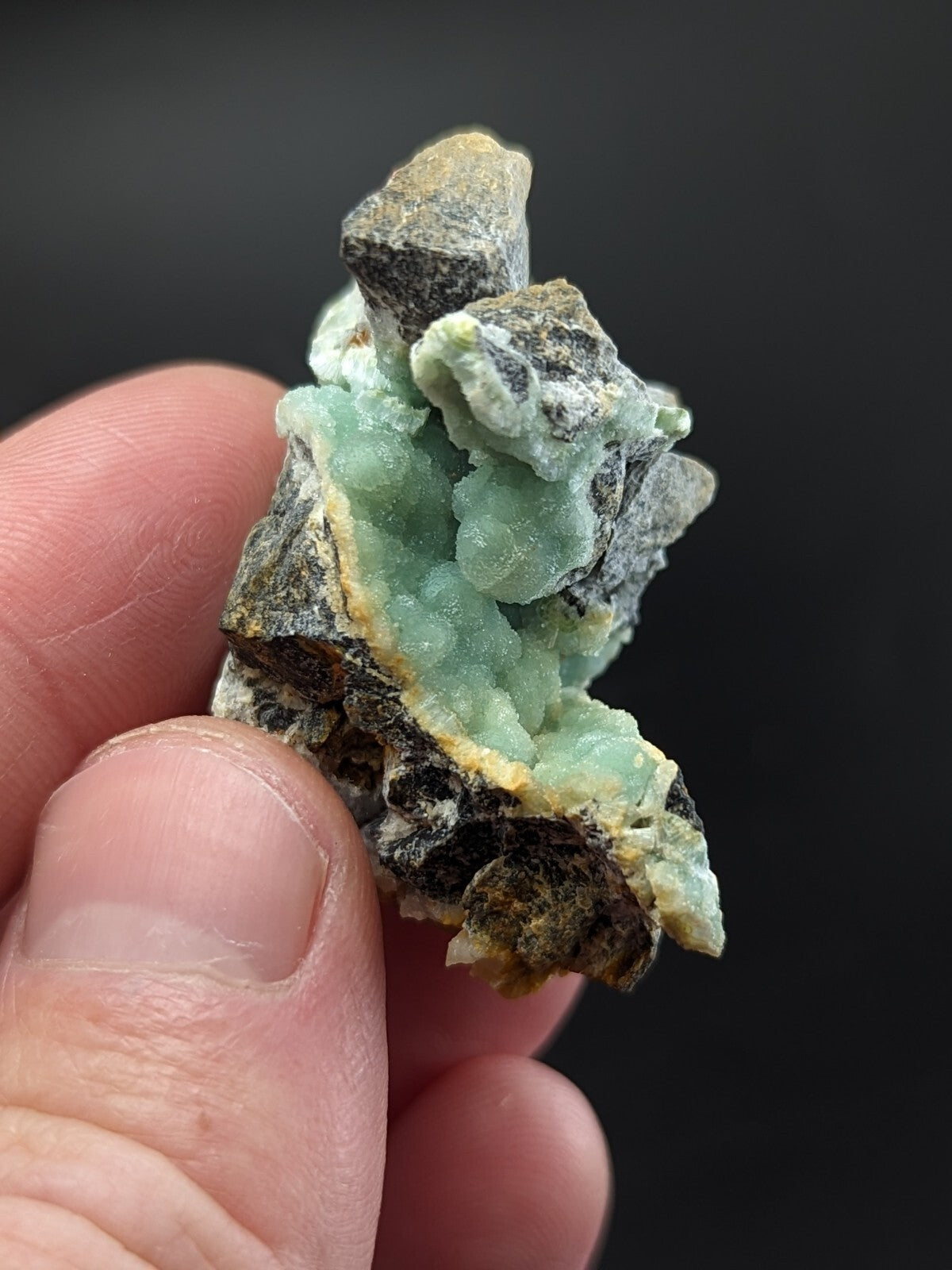 A hand holds a small, irregularly shaped mineral specimen featuring translucent light green crystals growing from dark gray and brown rock.