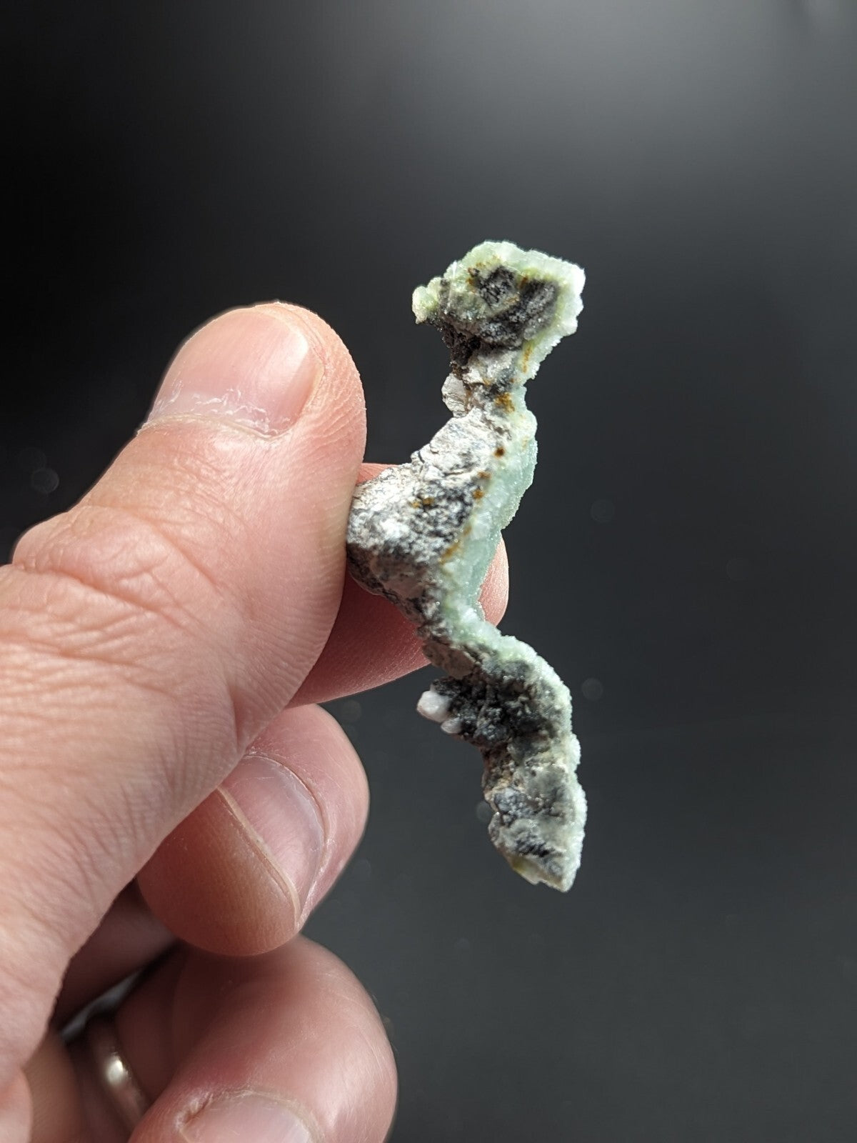 A hand holds a small, irregularly shaped piece of greenish-blue mineral with white and dark speckled patches.